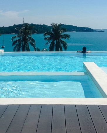 Luxury Resort. Woman Relaxing In Infinity Swimming Pool Water. Beautiful Happy Healthy Female Model Enjoying Summer Travel Vacation, Looking At Sea View. Summertime Recreation, Relax And Spa Concept.
